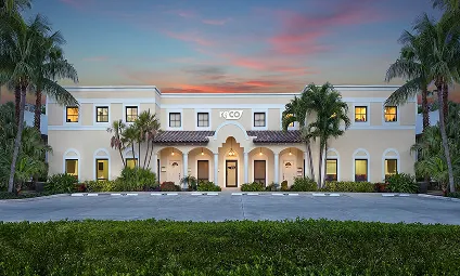 Front view of a two-story beige building with arched entrances and palm trees at sunset.