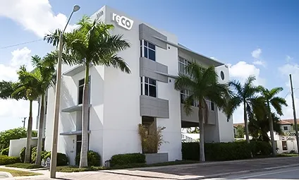 Modern white building with 'reco' signage, surrounded by palm trees under a blue sky.